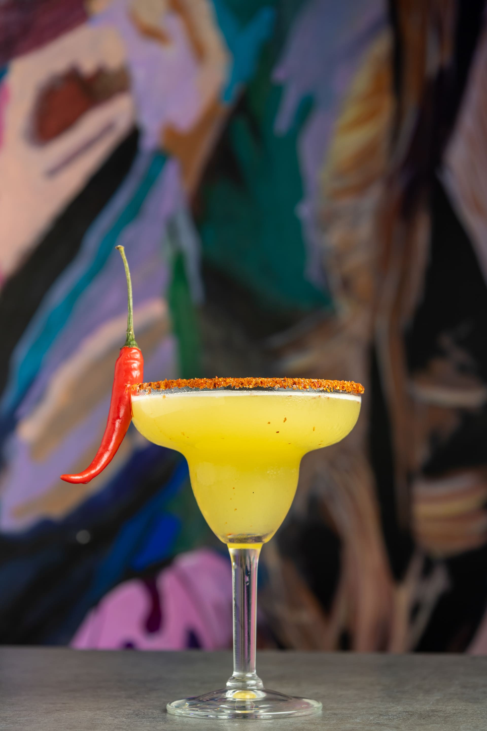 Margarita congelada con borde de chile y un ají rojo, sobre un mural de colores.